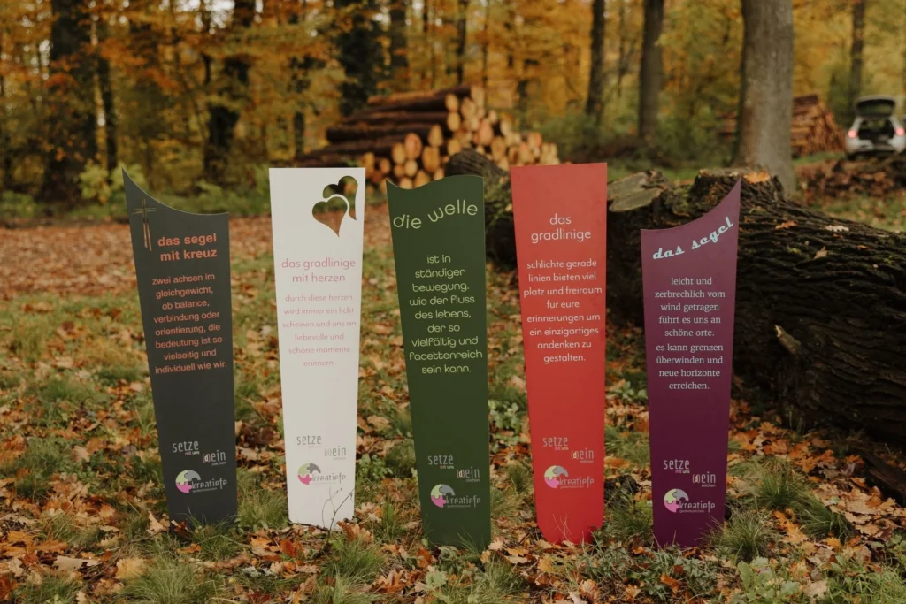 Fünf Gedenkzeichen von kreatiefe in verschiedenen Formen (Segel, Welle, gradlinig) und Farben, aufgestellt im herbstlichen Wald