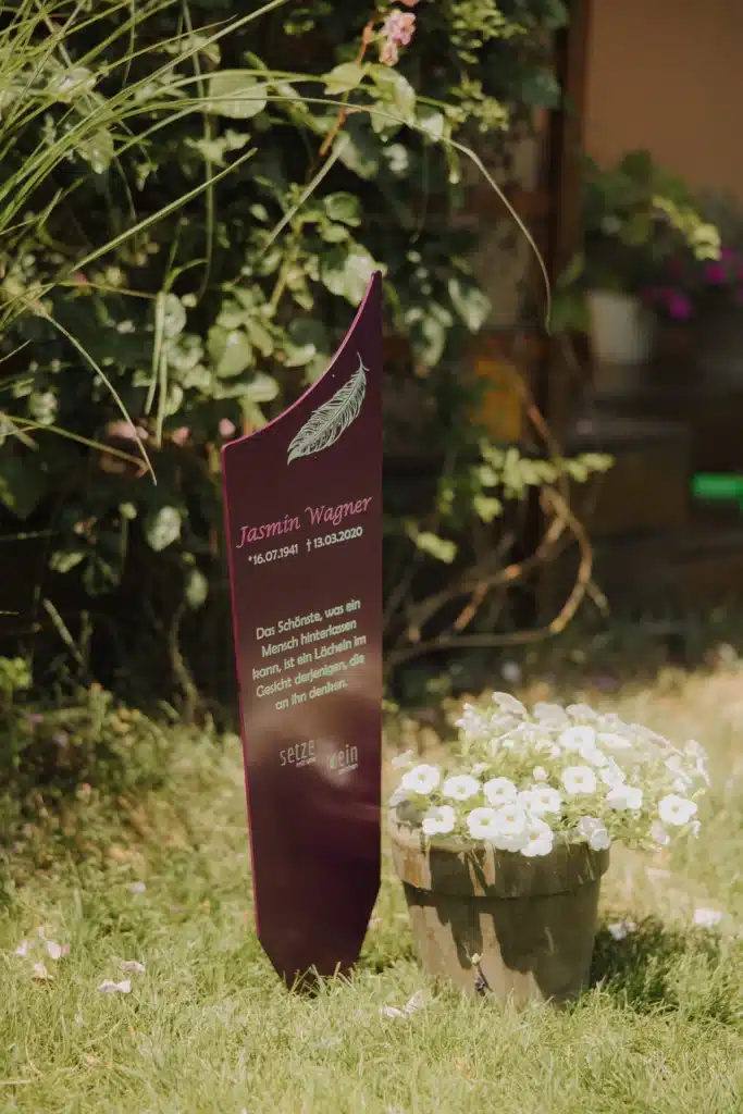 Individuelles Gedenkzeichen in Dunkelrot mit Feder-Motiv und persönlicher Inschrift, im sommerlichen Garten neben einem Topf mit weißen Blüten aufgestellt
